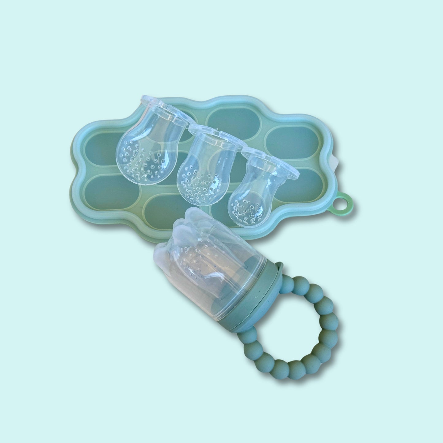Baby fruit feeder teether with beaded silicone handle and interchangeable silicone nipples, paired with a mint green freezer tray for prepping frozen fruit or puree portions